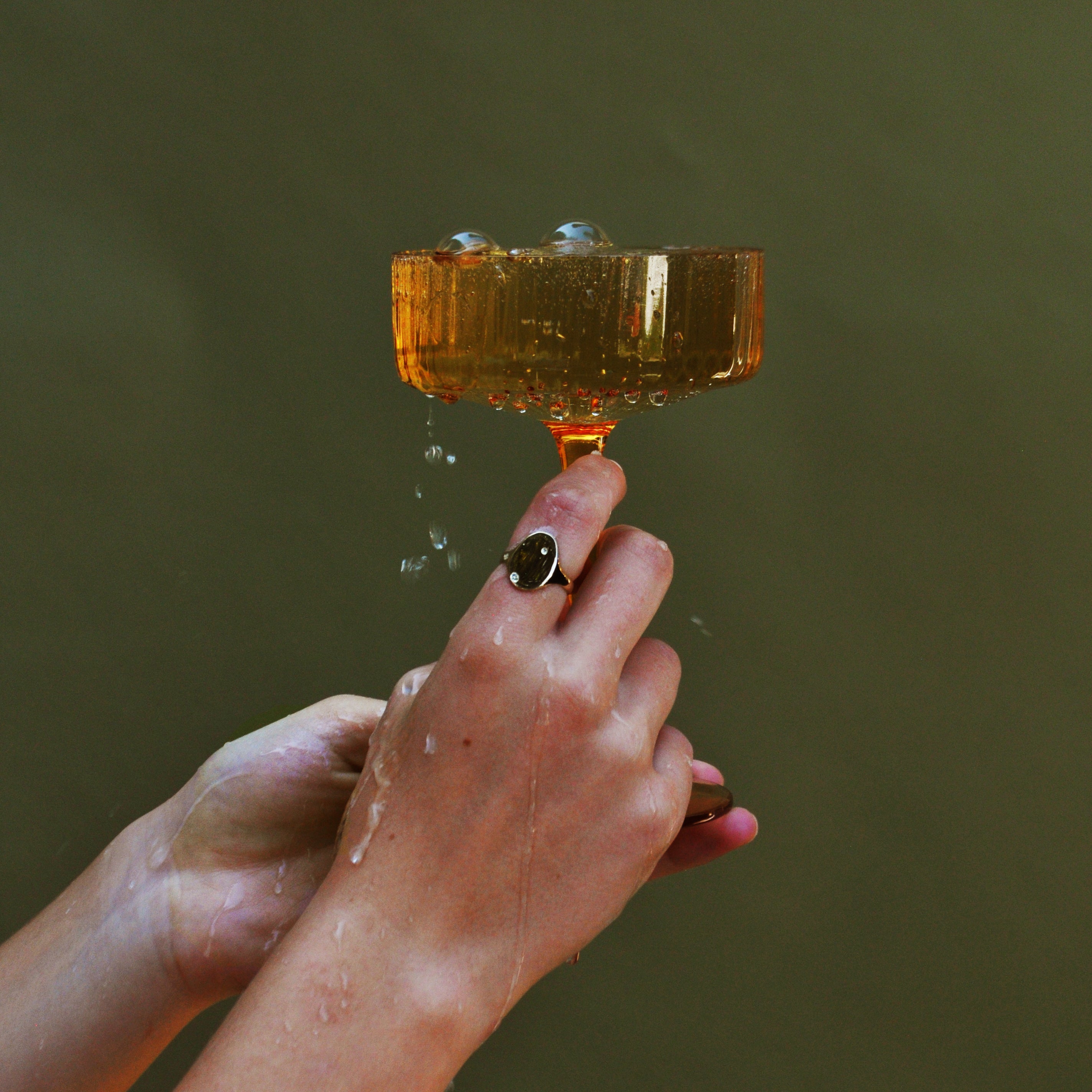 An image of a pair of hands holding a coupe glass with sparkling liquid overflowing wearing a gold signet ring on her index finger